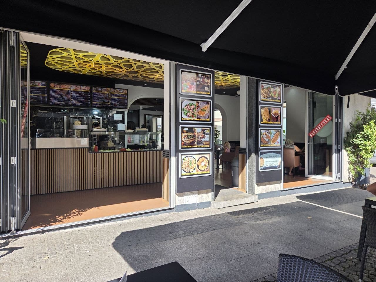 Das Miros Grill Haus von der Pfinztalstraße aus: geöffnete Falttüren, beleuchtete Speisentafeln an den Pfeilern und das goldene Deckengitter im Innenraum
